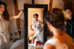 Une mariée en Charente qui termine son habillage en se regardant dans son miroir avec ses témoins à côtés. Tout le monde rigole et la mariée a une posture drole